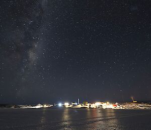 The Milky Way galaxy is visible in the night sky over a brightly lit station. There is a frozen over harbour in the foreground.