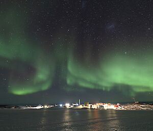 A bright green aurora with red and purple is in the night sky over a brightly lit station. There is a frozen over harbour in the foreground.