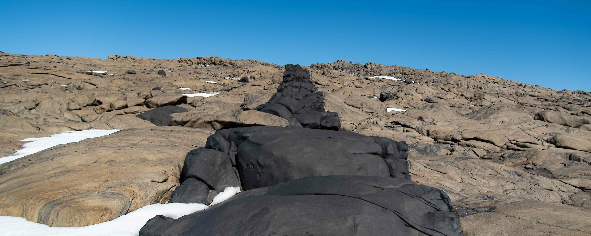 A line of black rock cuts through the surrounding beige rock of a hill with the blue sky visible above it.