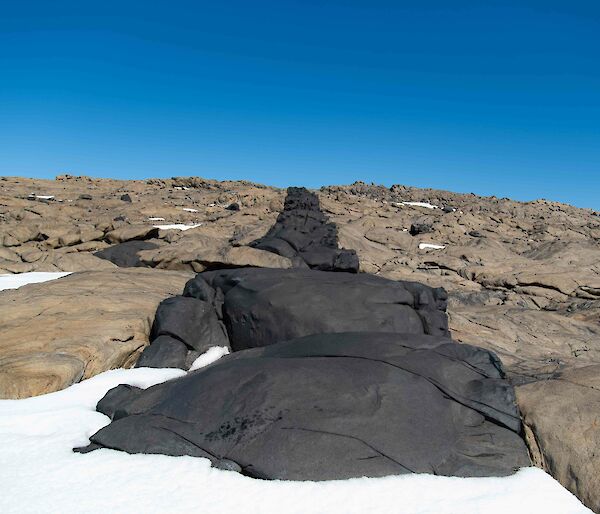 A line of black rock cuts through the surrounding beige rock of a hill with the blue sky visible above it.