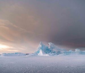Sharply scuplted triangular icebergs jut out of the flat sea-ice set against the backdrop of grey sun-streaked skies.