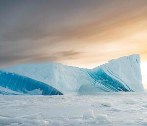 A white angular iceberg with three large jade-coloured streaks juts out of the sea-ice. The sky above is grey and golden.