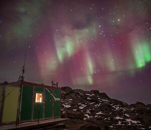 A star-studded night sky filled with ribbons of green and pink lights above a green field hut sitting in snowy, rocky hills.