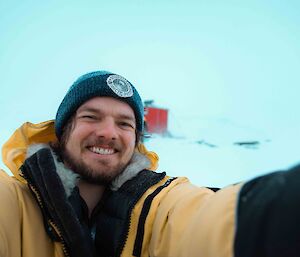 A smiling man takes a selfie while standing in the snow, a field hut visible behind him