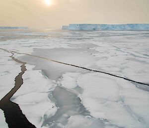 Cracked sea-ice in varying shades of white and silver with large icebergs visible in the distance. The sun can be seen trying to shine through a very grey sky.