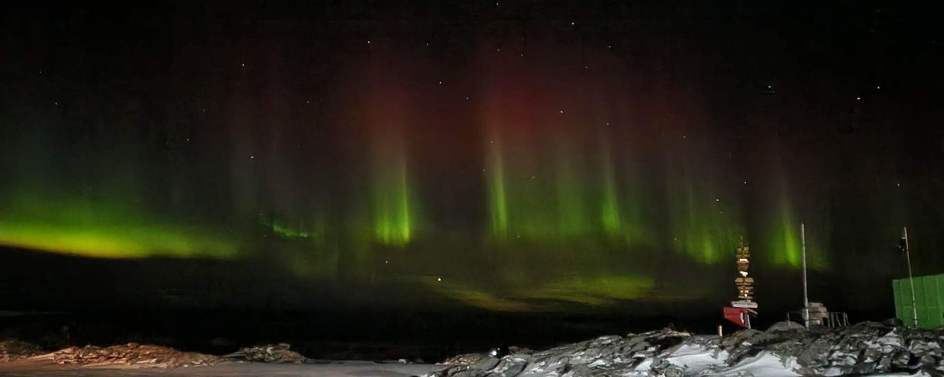 Green and red beams of light stretching across the dark sky. In the foreground is a rocky snow covered hill with a sign post on top, covered in lots of destination signs.