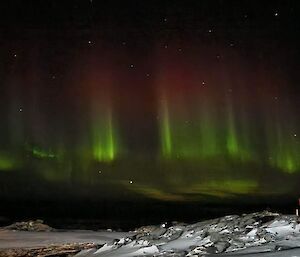 Green and red beams of light stretching across the dark sky. In the foreground is a rocky snow covered hill with a sign post on top, covered in lots of destination signs.