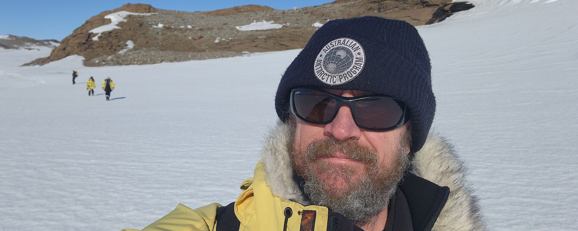 A man with a beard and wearing sunglasses is taking selfie with a snow covered ground behind him.