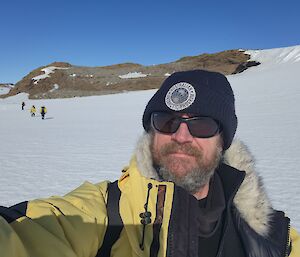 A man with a beard and wearing sunglasses is taking selfie with a snow covered ground behind him.