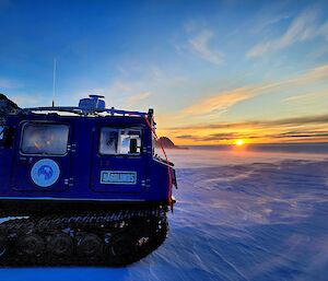 A blue Hägglands vehicle is to the left of frame with a wind swept ice covered plateau leading to the horizon with a setting sun.