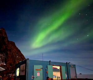 A bright green Aurora is visible in the night sky above a blue hut that is brightly lit inside. There is a rocky mountain slope in the rear left of frame.