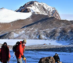 Three men are smiling at the camera on a rocky hillside. In the valley in the middle distance is a red hut and a blue Hägglunds vehicle. Another rocky mountain rises in the distance.