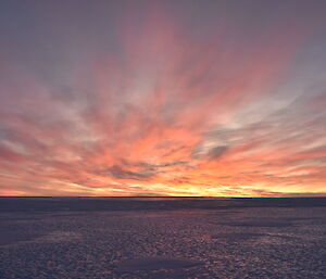 A orange and yellow glow from the sun below the horizon lights the sky and clouds. Ice extends to the horizon.