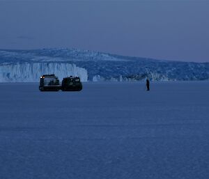 A Hägglunds vehicle is on sea ice with a person standing about 25metres in front of it. Ice cliffs are in the distance.