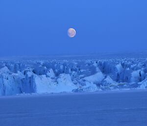 A nearly full moon can be seen clearly in the sky over the rough broken ice of a large glacier. Sea-ice is in the foreground.