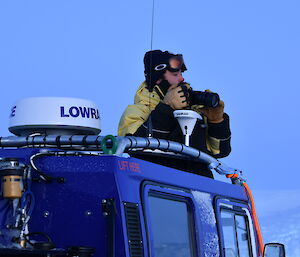 A man is holding a camera and looking into the distance. He is standing in the cupola of a blue Hägglunds vehicle.