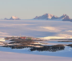 Brightly coloured station buildings and a wind turbine are visible in the distance on a rocky, snow covered landscape. It is surrounded by sea-ice and a rising ice plateau in the distance.