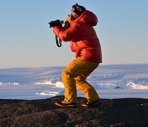 A man is taking a photo atop a rocky hillside. He is wearing a bright red jacket and yellow pants. He is holding a pair of yellow gloves in his mouth.