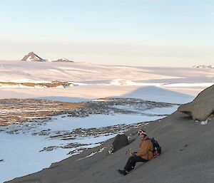 A man is sitting on a rocky slope looking at the camera. He is holding an ice axe. There is a vase ice covered landscape behind him with rocky mountains in the far distance.