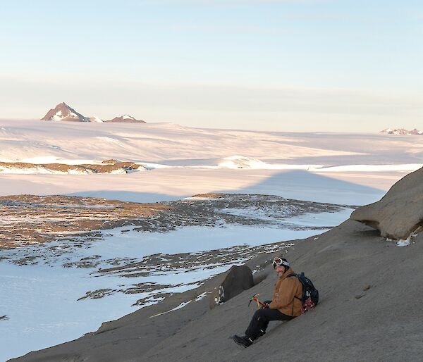 A man is sitting on a rocky slope looking at the camera. He is holding an ice axe. There is a vase ice covered landscape behind him with rocky mountains in the far distance.
