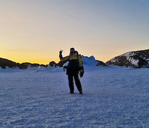 A woman is standing on the camera on the sea ice waving at the camera. There is a rocky island and small ice berg in the background and the sun is below the horizon.