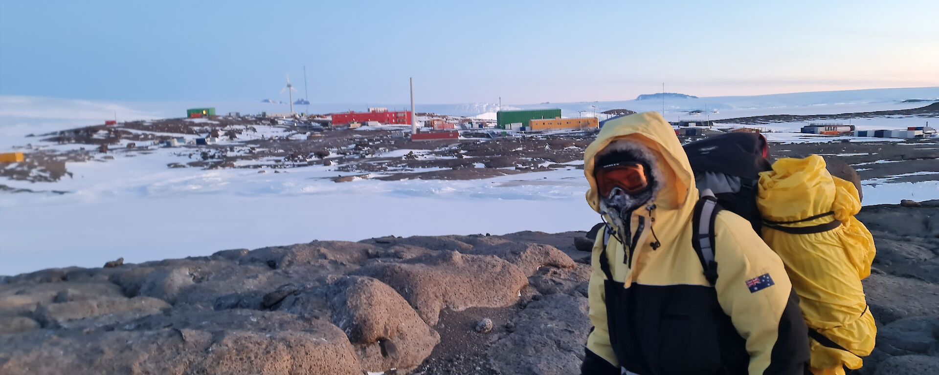 A man is looking at the camera. He is wearing goggles and heavy jacket. He is carrying a large backpack. Brightly coloured station buildings are in the background.