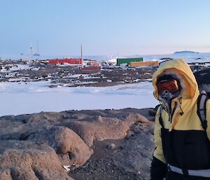 A man is looking at the camera. He is wearing goggles and heavy jacket. He is carrying a large backpack. Brightly coloured station buildings are in the background.