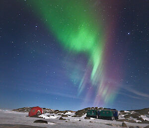 A bright green and purple aurora is visible in the night sky over a red dome hut, two Hägglunds vehicle and a larger hut which is just visible to the right of frame.