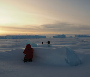 A woman wearing a bright red jacket is crouching behind a snow ridge and watching a number of Emperor Penguins in the distance. There are large ice bergs on the horizon and the sun is low in the sky.