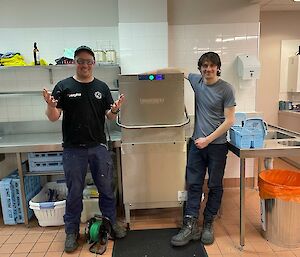 Two mean are smiling at the camera next to a commercial style Hobart dishwasher.