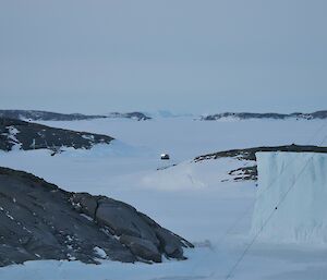A long distance photograph which shows a Hägglunds vehicle in the distance driving on the sea ice. There is a large ice berg and radio antenna to the right of frame and rocky islands are visible.