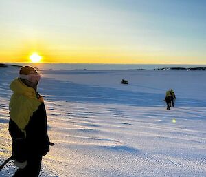 A woman is looking at the camera to the left of frame. She is wearing pink goggles and a heavy yellow and black jacket. Two men are in the background walking towards a Hägglunds vehicle that is parked out on the ice.