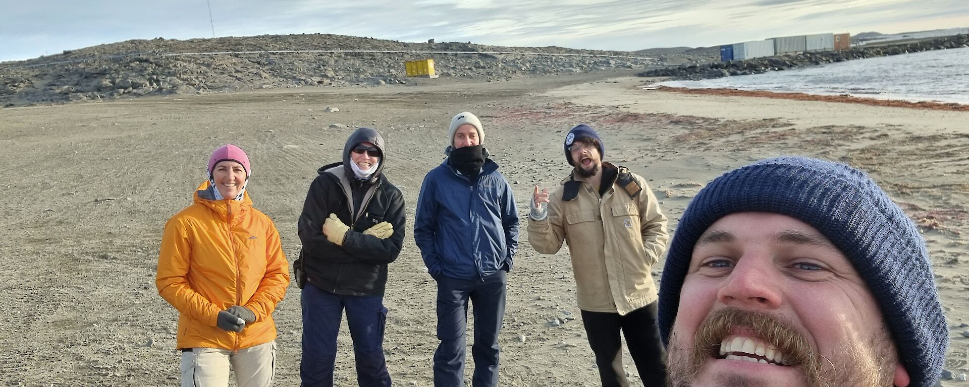 A group of five smiling people standing on a sandy beach in Antarctica with a hill in the background.
