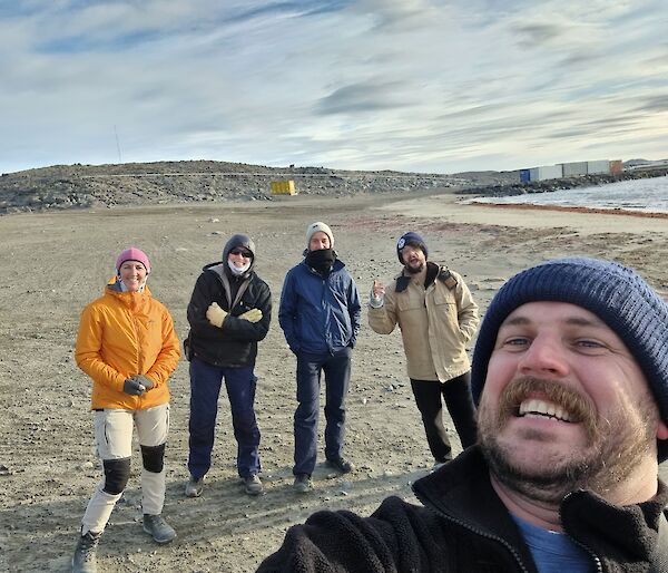 A group of 5 smiling people standing on a sandy beach in Antarctica with a hill in the background.