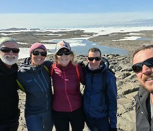 A group of five smiling people standing in a rocky Antarctic landscape with alternating areas of water, rocks and snow.