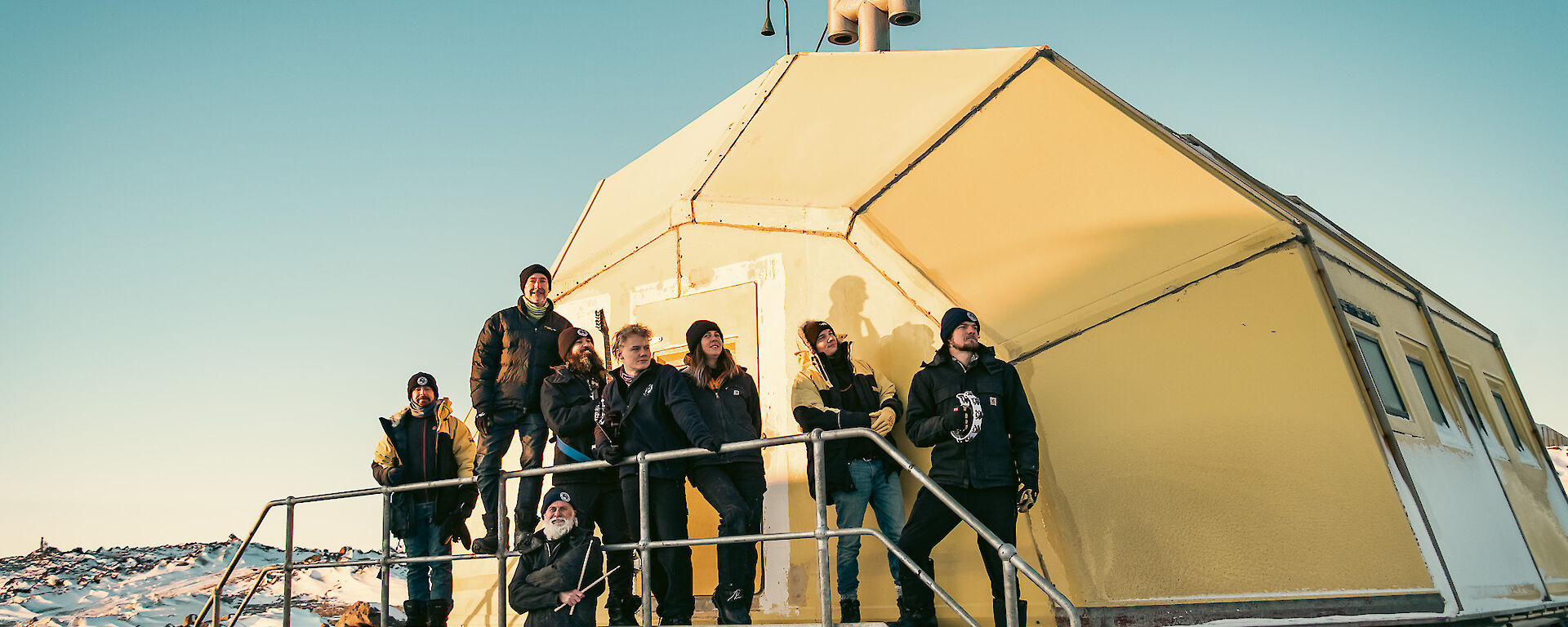 A group of eight people stand outside a yellow-hut that looks like a pineapple on its side.