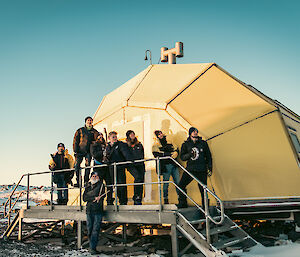 A group of eight people stand outside a yellow-hut that looks like a pineapple on its side.