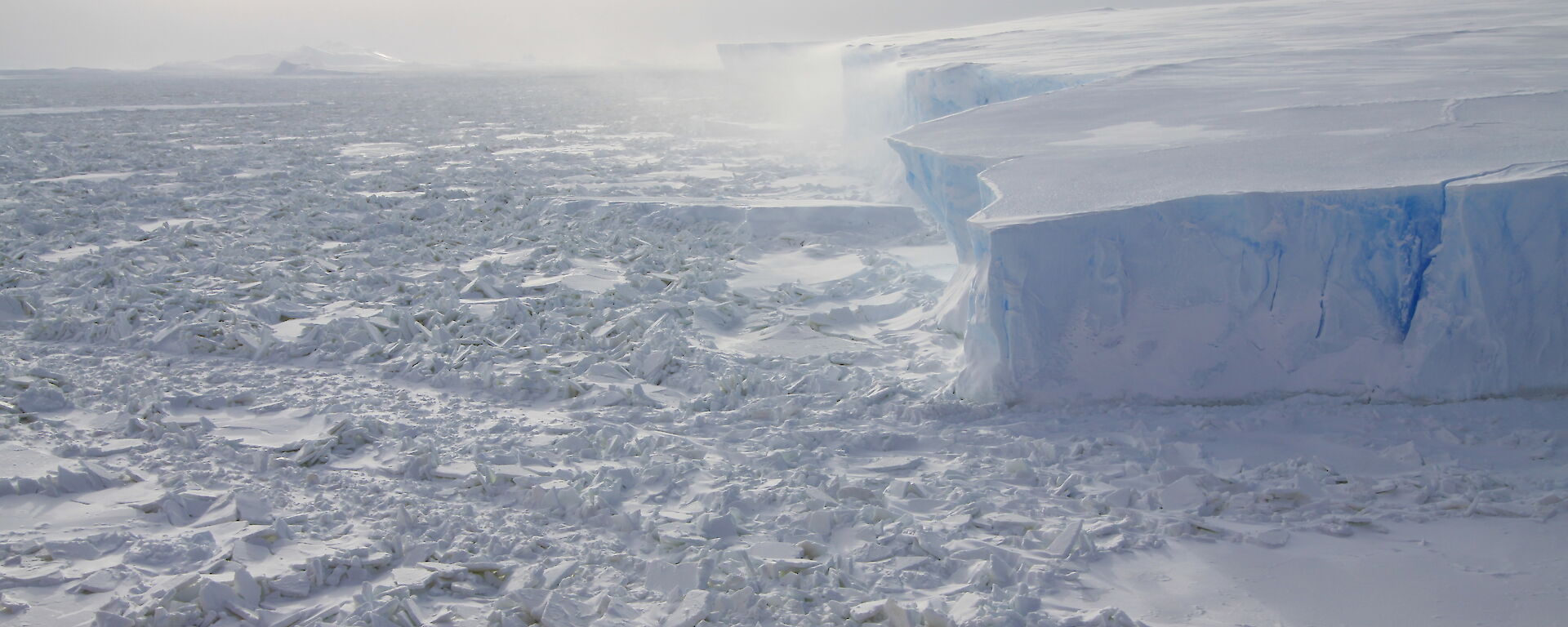 An icy plateau ending in ice cliffs to the right, over what looks like frozen sea and waves