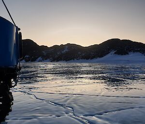 View from the side of a vehicle looking forward onto glassy, wet looking lake ice. Snow covered hills can be seen in the background.