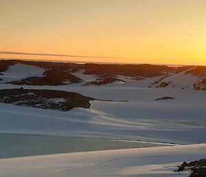 The view from the top of a hill showcasing snow covered rocky hills and an ice-lake in a valley.