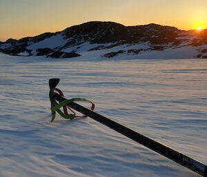 An ice-axe sits on the snow-covered surface of a frozen fjord with snow covered hills visible in the distance.