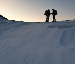 Two people wearing yellow snow clothing standing on top of a large mound of snow which is trailing off the edge of a hill.