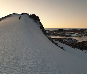 Two people in the distance climb up a large tail of snow that covers a rocky hill.