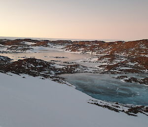 A view from the top of a snow-covered hill looking down onto frozen lakes surrounded by snow-covered rocky hills.
