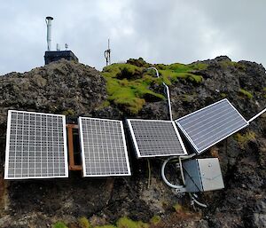 Solar panels and a grey box up against mossy rocks