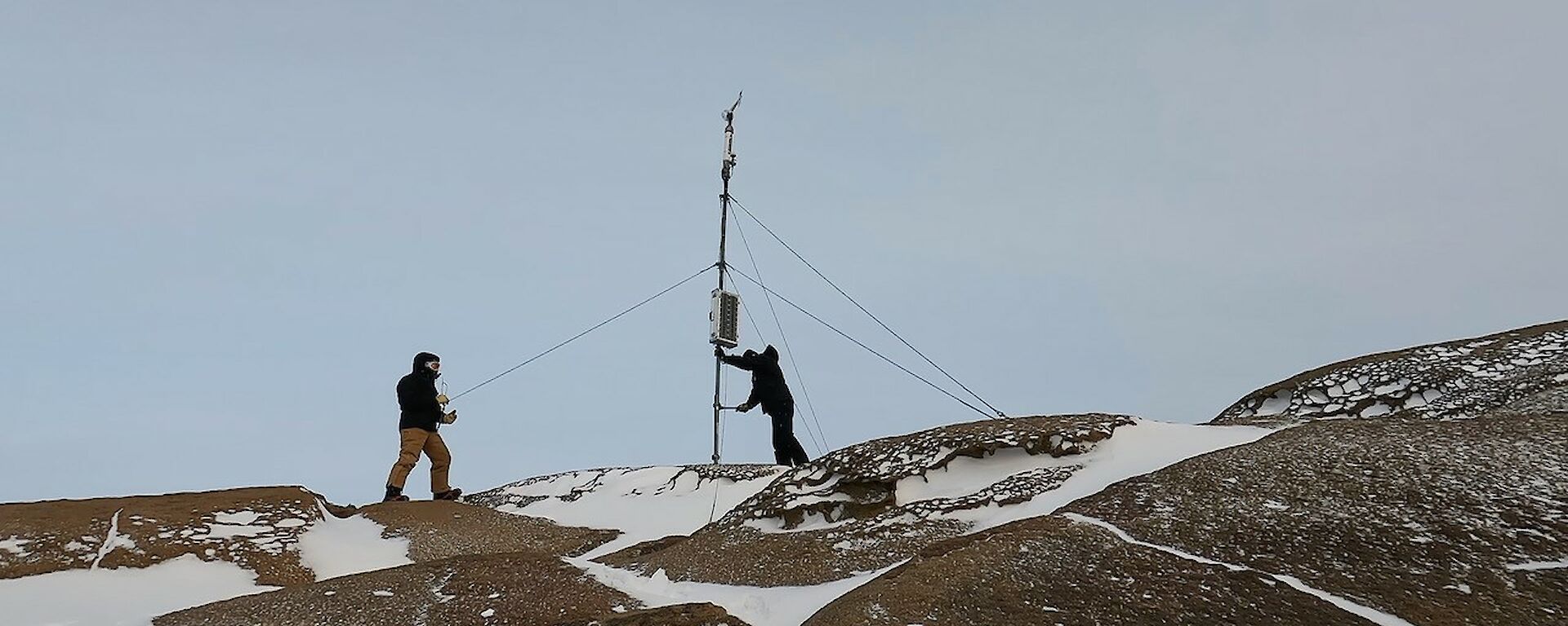 Two men stand on top of a rocky snow covered hill facing a mast. The mast holds a wind vane on top of it.
