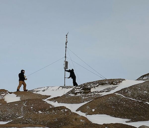 Two men stand on top of a rocky snow covered hill facing a mast. The mast holds a wind vane on top of it.