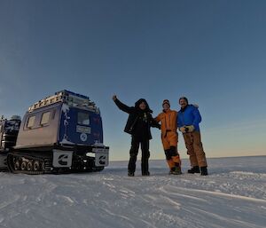 Three people stand facing the camera with arms around each other. To the left of the photo is a Hagglund tracked vehicle. The ground is the flat icy surface of the Peterson Glacier.