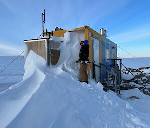 A man stands on a large pile of snow, at the end of a shipping container building. He is holding a shovel and the end of the building is half buried in the snow.