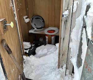 This photo shows an open toilet door, and the remenants of snow inside the toilet! There is a shovel leaning against the toilet door. The toilet itself is a bucket placed under a wooden board.